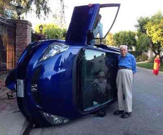 Un homme âgé posant à côté de leur voiture retournée, alors que sa femme est encore coincée à l'intérieur.